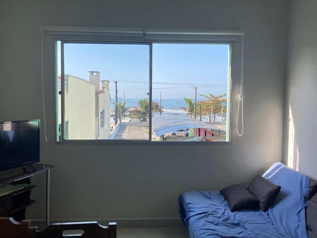 Sala do Apto vista p o mar Ubatuba. Do lado direito um sofá com almofadas, do lado esquerdo uma raque com televisão, no meio uma janela com vista para a praia. Foto para ilustrar post sobre airbnb na Praia Grande.