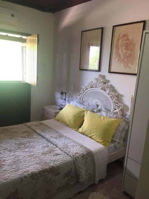 Foto do quarto em Apartamentos Aconchegantes, que ilustra o post de airbnb na Praia do Forte. Há uma cama de casal com cabeceira branca ornamentada. Na esquerda há uma mesa de cabeceira com abajur e janela.