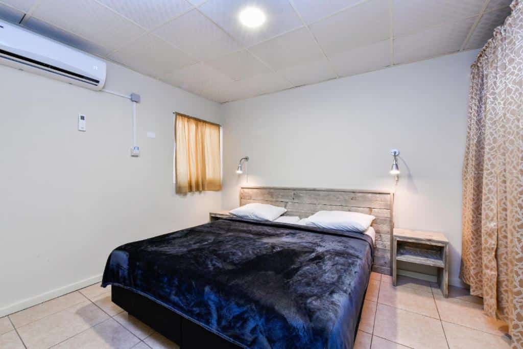 Quarto do Aruba Blue Village Hotel and Apartments com uma cama de casal no centro da suíte com cabeceira de madeira e dois abajures em cada lado da cama. No canto superior esquerdo é possível ver um ar-condicionado. Ele é um dos hotéis baratos em Aruba.
