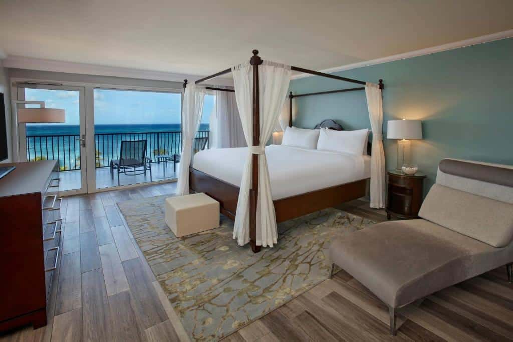 Quarto do Marriott Resort & Stellaris Casino. Uma poltrona e uma cama de casal do lado direito com uma luminária de cada lado. Do lado esquerdo uma cômoda com televisão. No fundo, a varanda com vista para o mar. Foto para ilustrar post de onde ficar em Aruba.