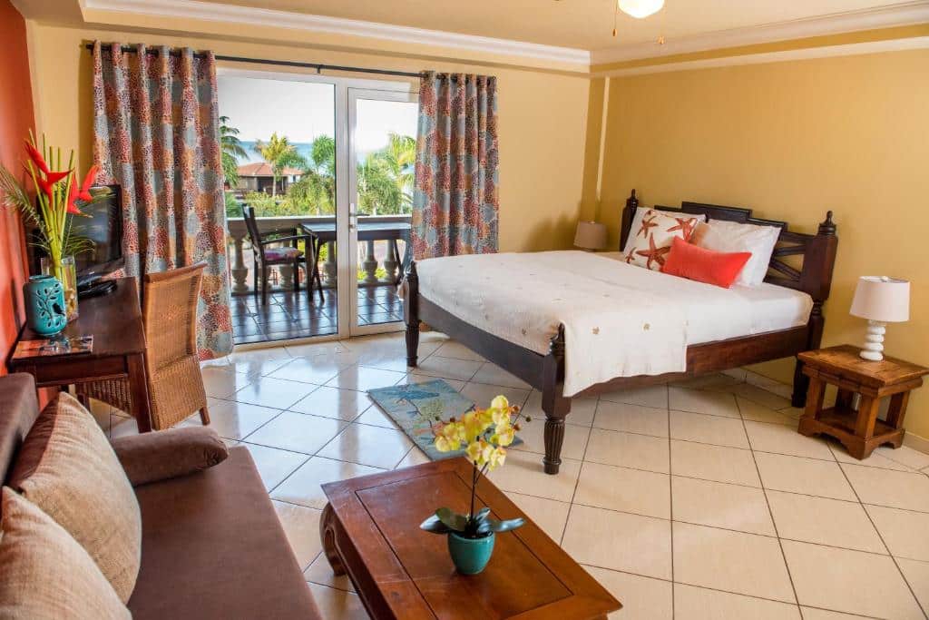 Quarto do Aruba Surfside Marina com uma cama de casal no lado direito da imagem e móveis de madeira no lado esquerdo, como uma mesa de centro, uma mesa de escritório e um sofá encostado na parede. Ao fundo há duas portas de vidro que dão acesso a varanda particular e a vista para o mar. A imagem mostra um dos hotéis baratos em Aruba.
