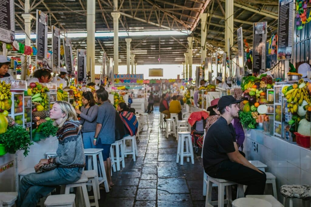 mercado de san pedro, em cusco, com várias barracas de frutas e verduras, pessoas sentadas em banquinhos brancos e atendentes em pé.