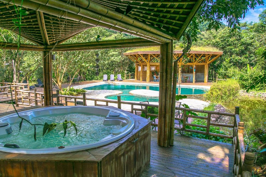 Parte do Banana Bamboo Ecolodge que mostra em primeiro plano uma banheira de hidromassagem coberta por uma cobertura de bamboo. Ao fundo tem uma piscina ao ar livre e um espaço coberto e muitas árvores verdes em volta. Imagem ilustrando post Airbnb em Picinguaba.