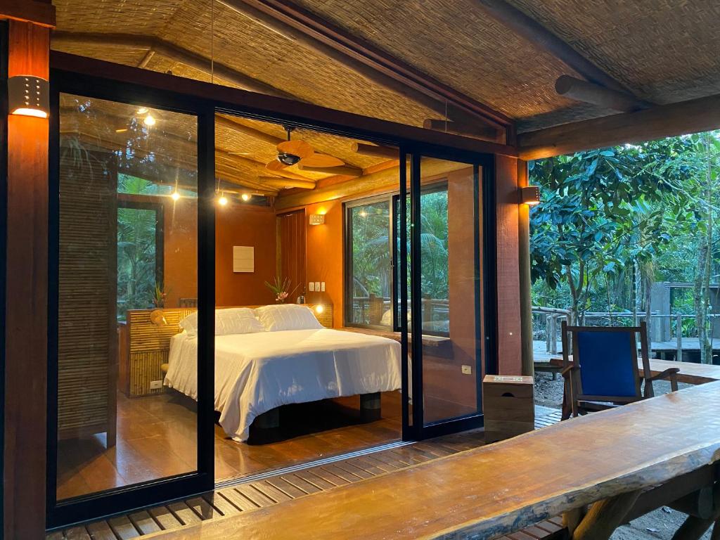 Quarto do chalé do Banana Bamboo Ecolodge que mostra uma cama de casal em ambiente com luz amarela dentro da construção do chalé que tem portas e janelas de vidro, chão de madeira e varanda com cadeira para ver a natureza. Imagem ilustrando post Airbnb em Maranduba.