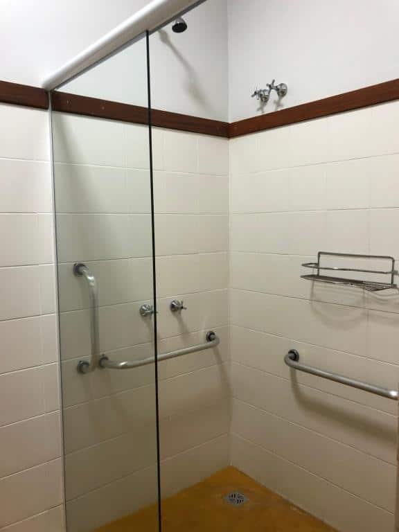 Box do banheiro da Pousada Altamira. Na parede do lado direito há uma barra de apoio e na parede do lado direito há duas barras de apoio em baixo do registro do chuveiro. No lado direito há uma porta de vidro. Imagem utilizada para ilustrar o post pousadas pet friendly em Ilhabela.