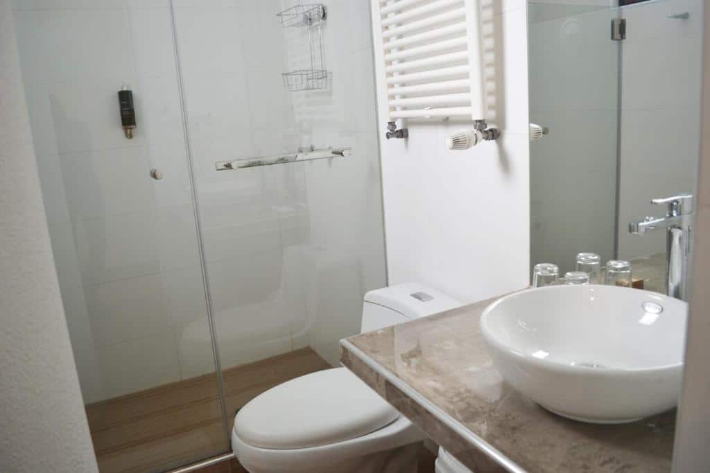 banheiro do Katari Apart Hotel at Qorikancha com um box de vidro no chuveiro, um porta toalhas aquecedo preso a parede em cima do vaso sanitário e uma pia com quatro copos de vidro.
