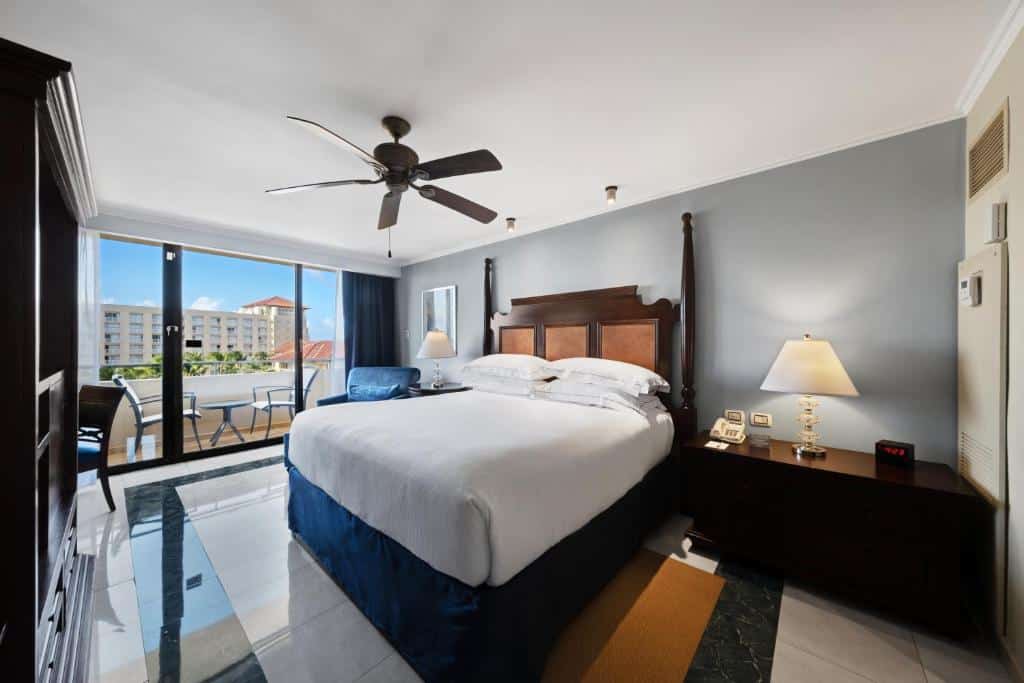 Quarto do Barceló Aruba - All Inclusive. Uma cama de casal no meio, de cada lado um abajur e uma cômoda. Em cima um ventilador de teto, no fundo uma poltrona e a varanda com cadeiras.