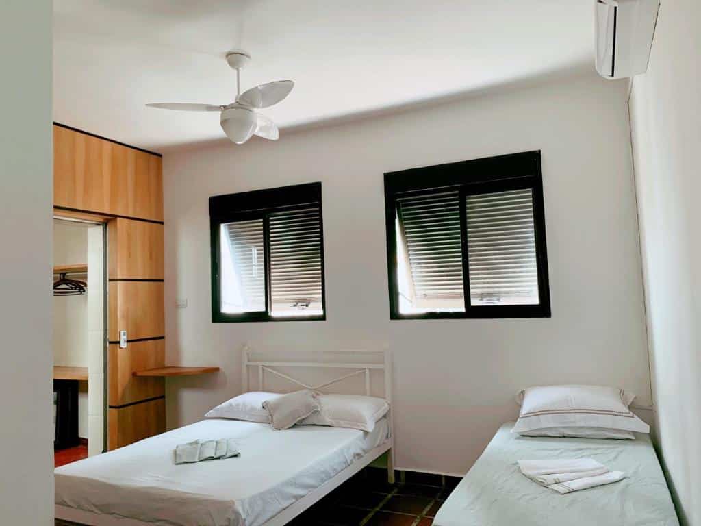 Um quarto na BEACH HOUSE Unidade 1 Casa de Praia Guarujá. Há uma cama de casal na esquerda e outra de solteiro na direita. Na parede atrás há duas janelas.