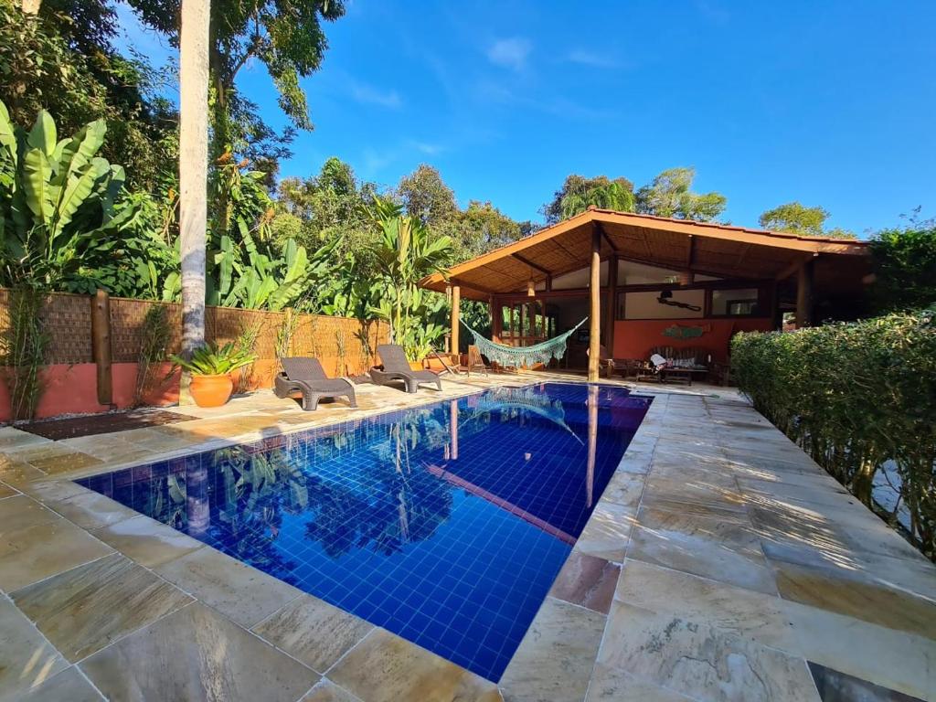 Piscina da Beach House Prumirim. Ela é retngular e está no meio de uma área aberta. Ao seu lado estão duas espreguiçadeiras. Ao fundo há uma rede e a entrada da casa.