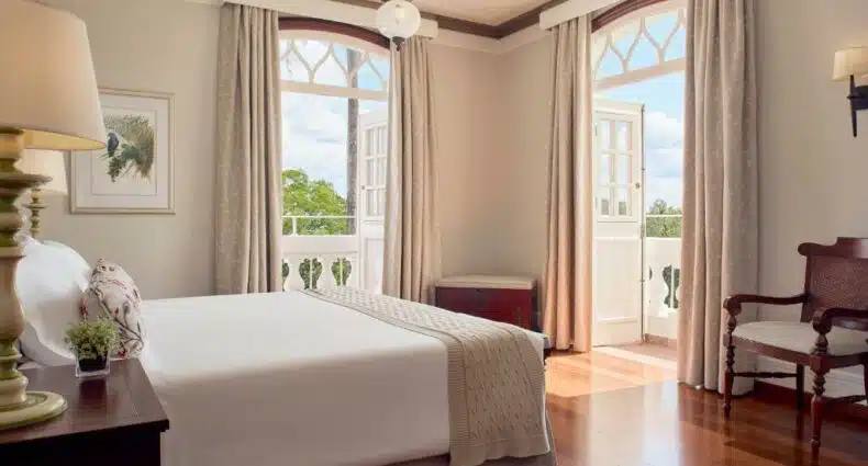 Quarto no Belmond Hotel das Cataratas, mostrando uma cama de casal na esquerda, que possui uma mesa de cabeceira com abajur em cima do seu lado. Na frente da cama há duas janelas com varanda que possuem vista para as cataras. Há uma poltrona no canto inferior direito.