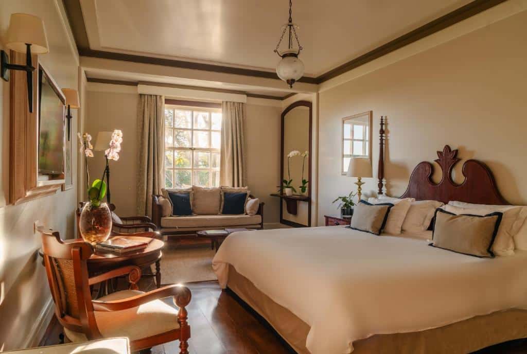 Uma suíte no Belmond Hotel das Cataratas. Há uma cama box de casal na direita, e na frente dela se vê uma poltrona e uma mesa redonda com vaso de flores e um livro. No fundo do quarto há uma janela, um sofá e um espelho alto.