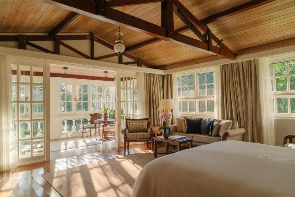 Foto do quarto do Belmond Hotel das Cataratas, ilustrando o post sobre Onde ficar em Foz do Iguaçu. O espaço é grande, há uma cama box de casal no canto inferior direito. Na frente da cama há um sofá com poltrona e mesa de centro. Já mais ao fundo há uma porta de vidro com outro espaço de descanso que dá acesso para a varanda da suíte.