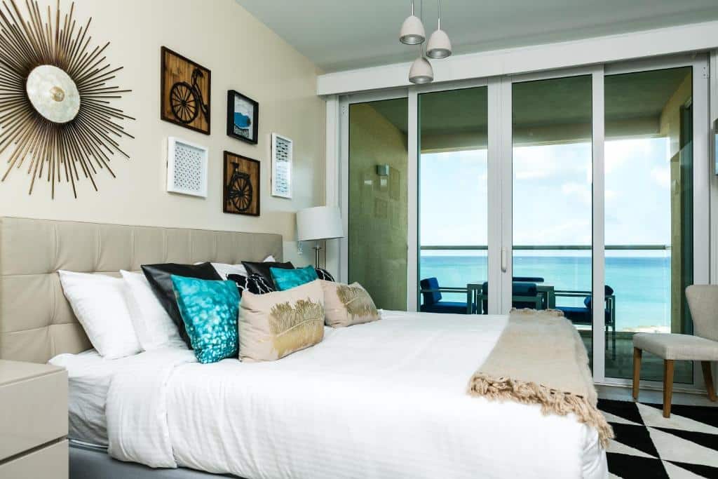 Quarto do Blue Residences. Uma cama de casal está no meio, ao lado de uma porta de vidro que leva até a varanda. Nela está uma mesa com cadeiras de frente para o mar.