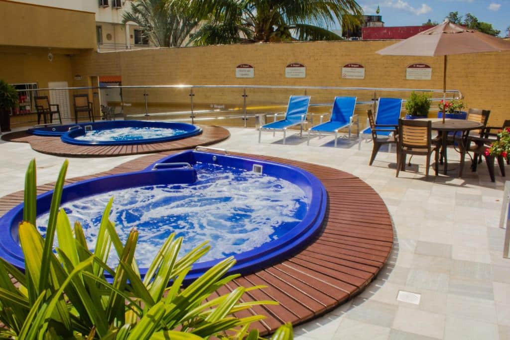Parte do Bogari Hotel que mostra duas piscinas redondas azuis, uma planta, três espreguiçadeiras e uma mesa com quatro cadeiras e um guarda-sol. Imagem ilustrando post Hotéis em Foz do Iguaçu.