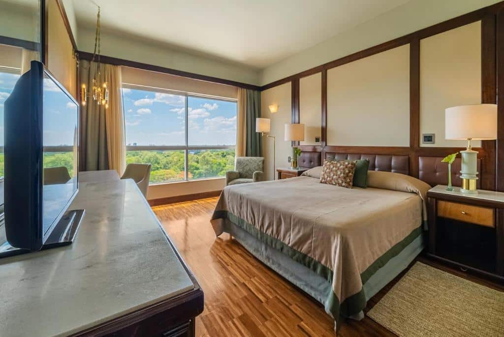 Foto do quarto do Bourbon Cataratas do Iguaçu Thermas Eco Resort, ilustrando o post sobre Onde ficar em Foz do Iguaçu. Há uma cama box de casal na direita, e dos dois lados dela há uma mesa de cabeceira com abajur. No fundo do quarto há uma janela com vista. Na frente da cama há uma cômoda com TV.