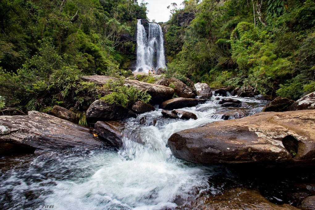 Queda-d'água da Cachoeira dos Garcias em Aiuruoca formando um poço quando chega no chão e em volta várias árvores verdes e montanhas ao fundo durante o dia. Ilustrando post airbnb em Aiuruoca.