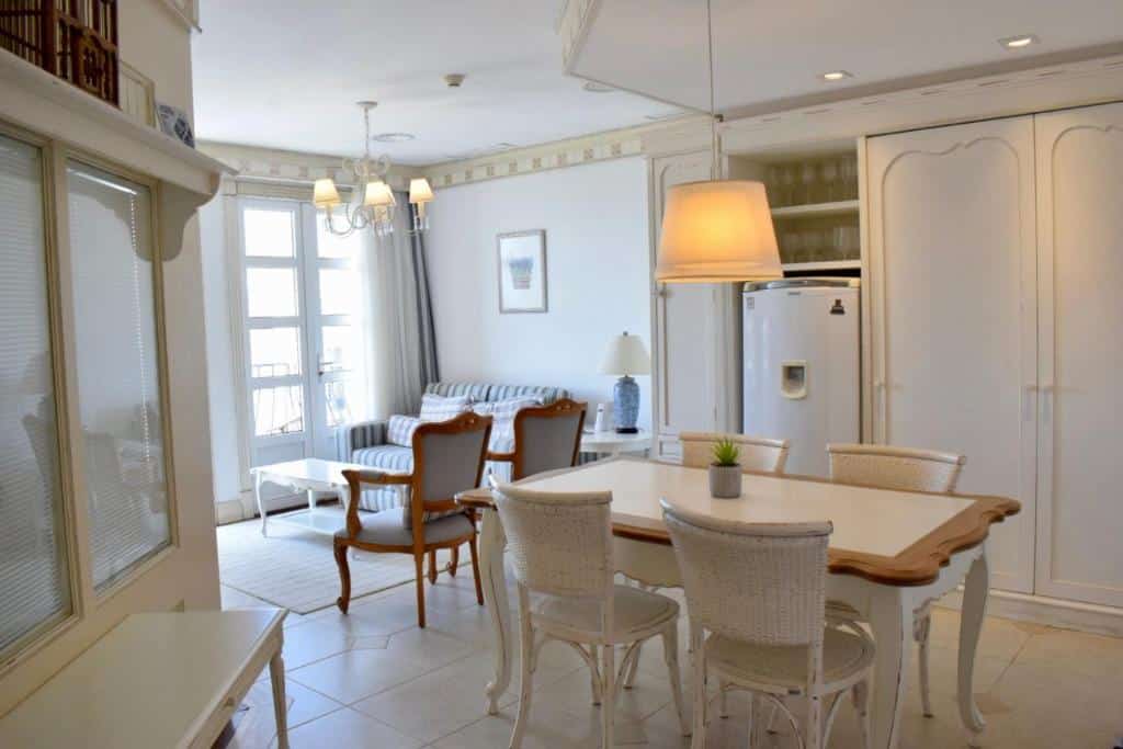 Sala de estar do airbnb Il Campanario - Cobertura. No lado direito da imagem há uma mesa com quatro lugares, na parede a frente da mesa há um armário e uma geladeira ao lado. No lado esquerdo ao fundo é possível ver a sala, que têm duas cadeiras, um sofá e uma porta com cortinas.