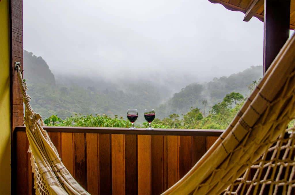 Vista da varanda no Cantinho Pedra da Gávea. Há uma rede e duas taças de vinho apoiadas no parapeito da janela. A vista é para as montanhas.