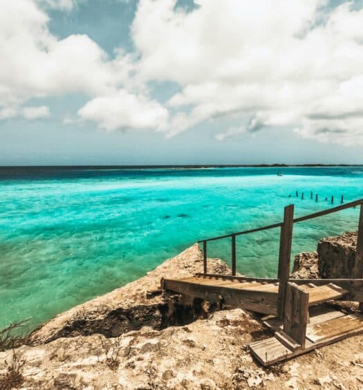 Imagem durante o dia uma ponte de madeira em Aruba que dá acesso ao mar do lado direito da imagem com vista para o mar esverdeado. Representa pontos turísticos em Aruba.