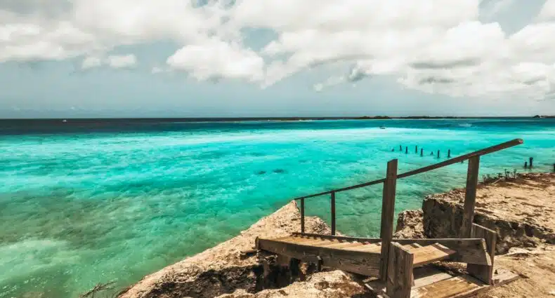 Imagem durante o dia uma ponte de madeira em Aruba que dá acesso ao mar do lado direito da imagem com vista para o mar esverdeado. Representa pontos turísticos em Aruba.