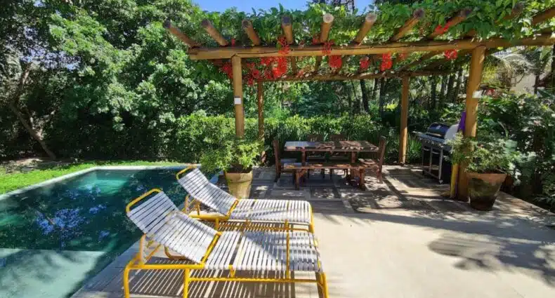 Imagem da área de lazer da Linda Casa Praia do Pauba em condomínio durante o dia com piscina do lado esquerdo da imagem e do lado direito duas cadeiras. Representa airbnb em São Sebastião.