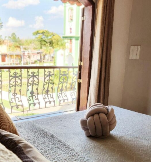 Quarto do PousaDinháChica Hospedagem com cama de casal do lado esquerdo da imagem e do lado esquerdo da cama uma porta com acesso a varanda. Representa aibnb em São João del Rei.