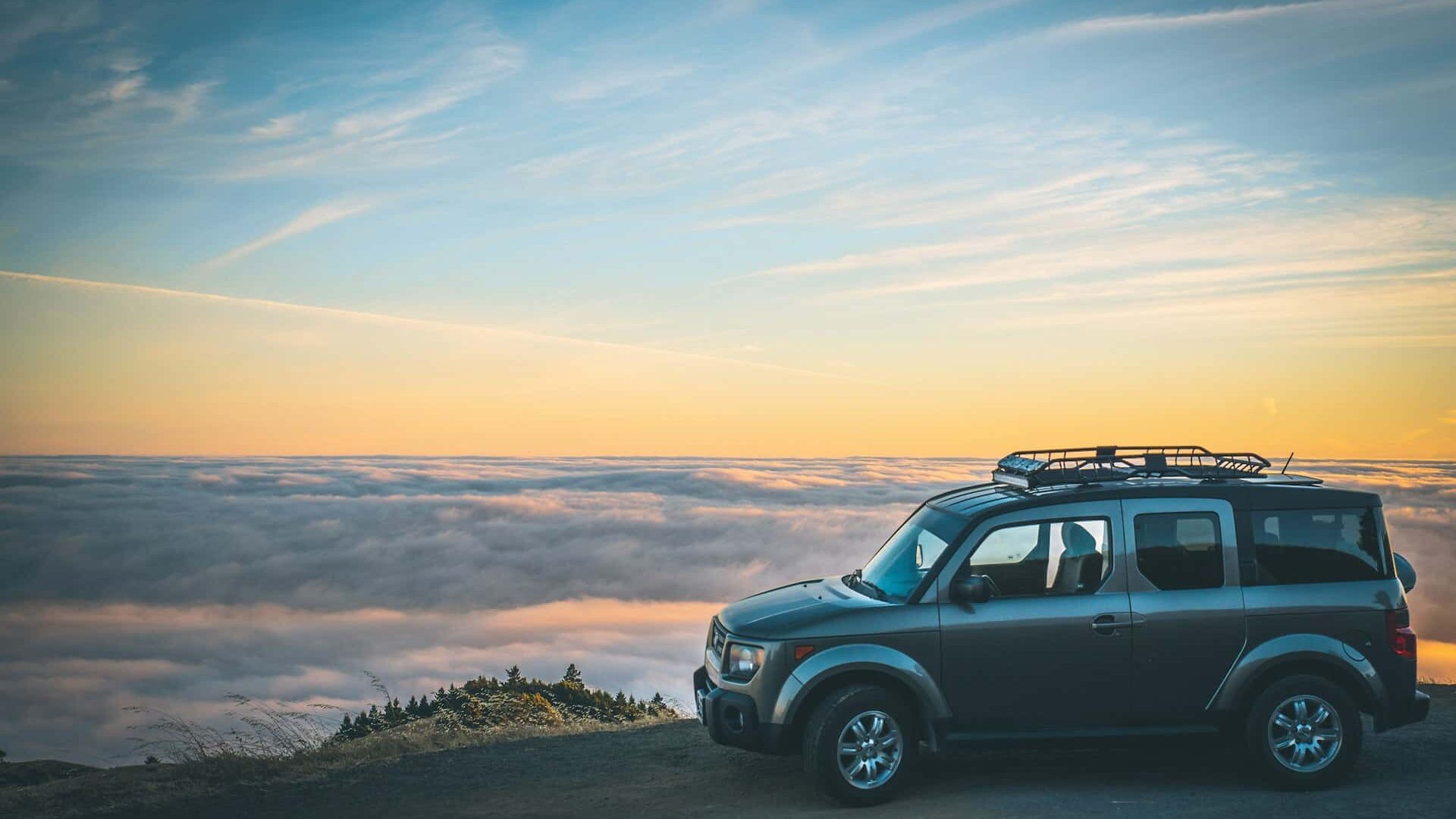 Carro estacionado em um mirante com núvens no horizonte e o céu com o pôr do sol. Foto de Casey Horner via Unsplash