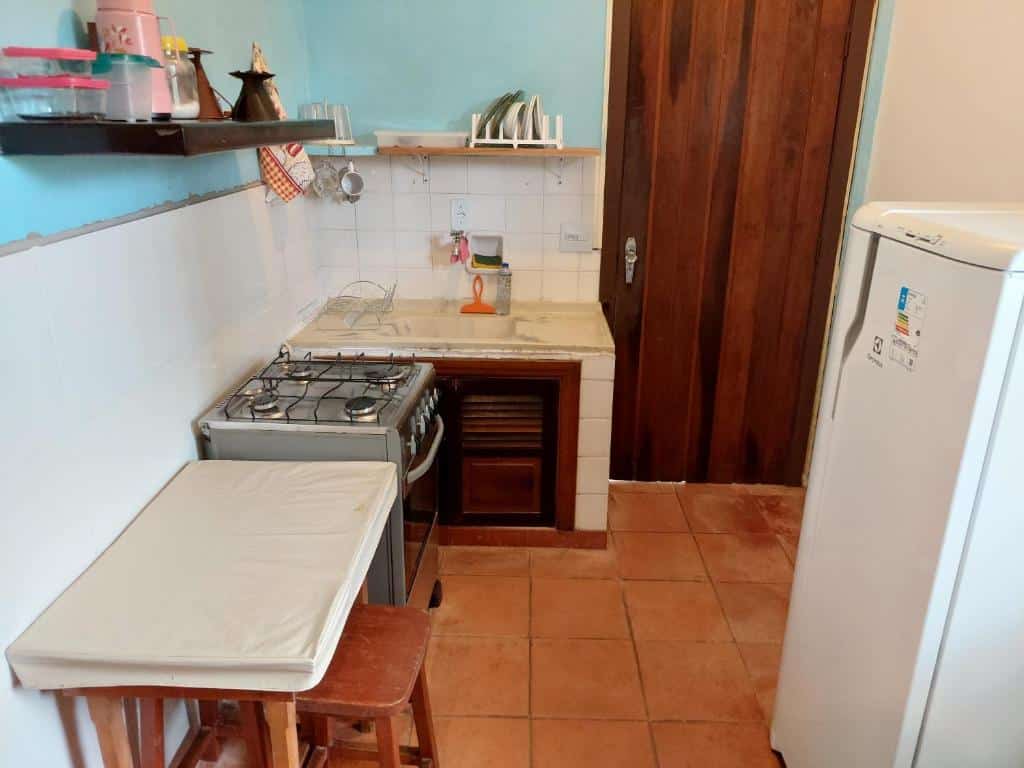 Cozinha do Casa a 130 metros da praia de Boiçucanga. Uma geladeira está de frente para uma pequena mesinha. Ao lado dessa mesa está um fogão e logo em seguida uma pia. Na parede duas existem duas prateleiras com utensílios.