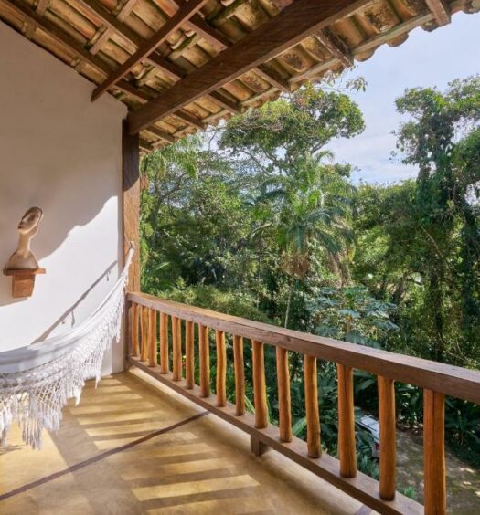 Varanda de um dos quartos da Casa Amaargi Picinguaba que mostra do lado esquerdo uma rede e do lado direito a paisagem de árvores verdes. Imagem ilustrando post Airbnb em Picinguaba.