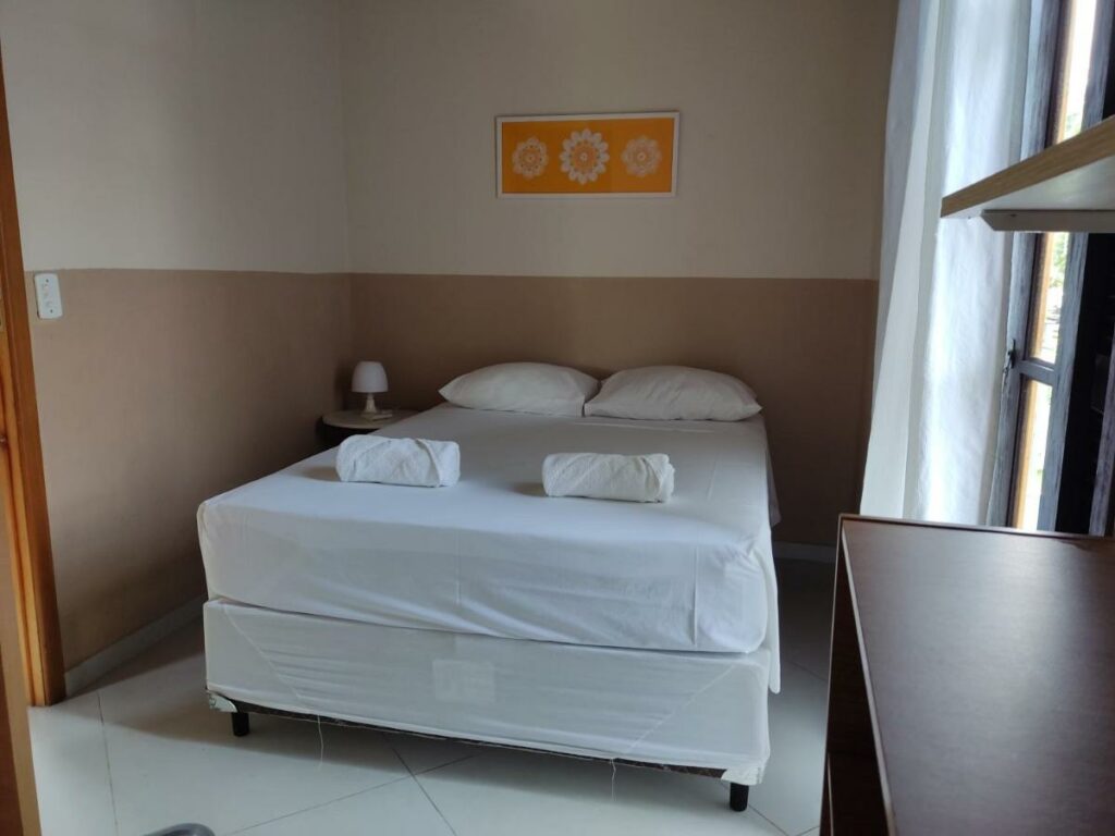 Quarto da Casa Azul Perequê, um dos airbnb em Ilhabela. Uma cama de casal com mesinha de cabeceira e abajur do lado esquerdo e toalhas dobradas aos pés está encostada na parede central, e na lateral direito está uma janela e uma cômoda.