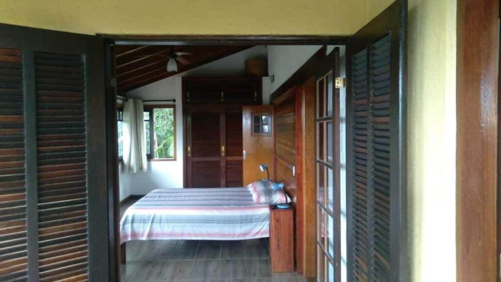 Quarto da Casa cachoeira praia Prumirim Ubatuba. Uma cama de casal está no meio, ao lado de um armário de madeira. Do seu outro lado está uma porta.