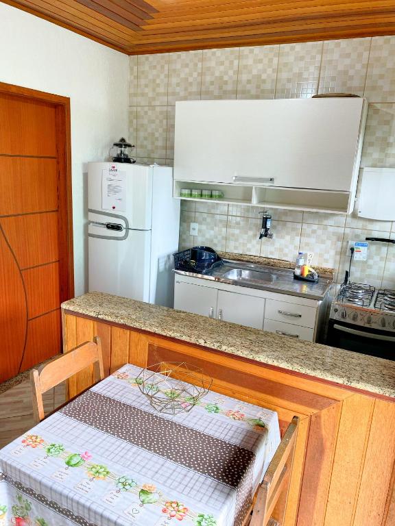 Cozinha na Casa da Lilyan. Há uma mesa com cadeiras encostada do lado e uma bancada na cozinha, essa que possui armários, uma geladeira, pia e fogão.