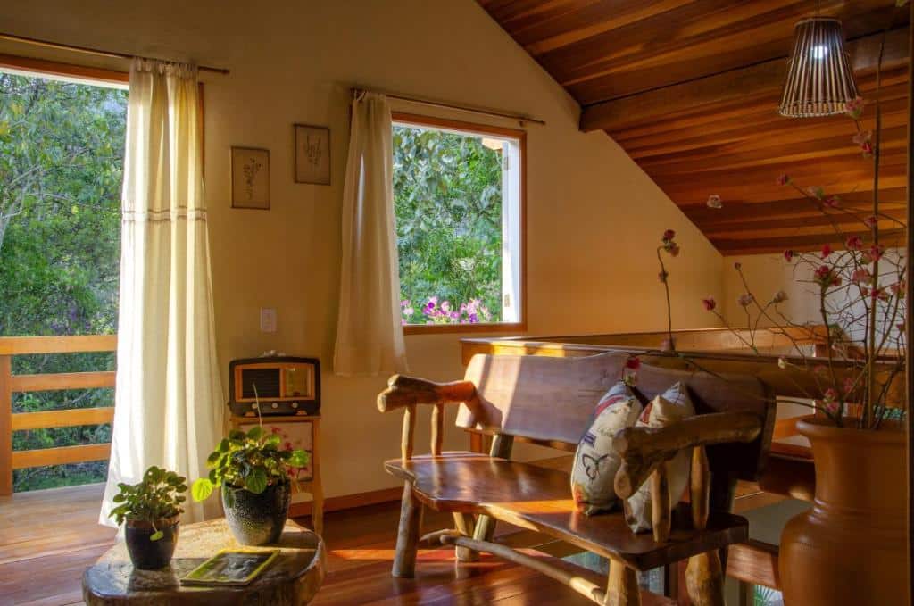 Sala na Casa de Férias e Hospedagem Ana Lua. É um espaço com varanda e janelas. O piso é de madeira, há um banco de madeira na direita com almofadas, uma mesa de centro com plantas e um rádio.