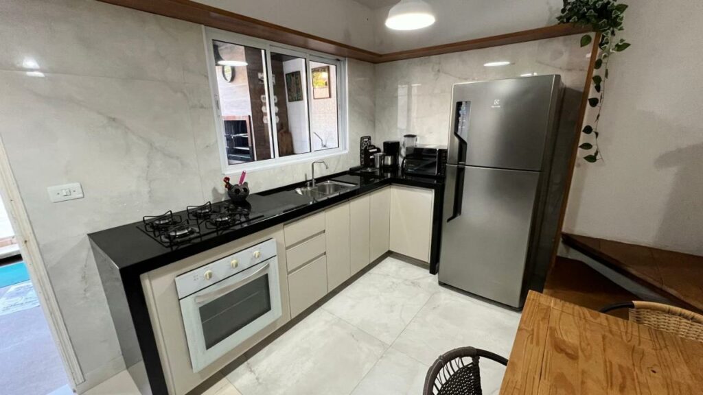 Cozinha da Casa de Praia com piscina. No lado esquerdo o fogão estão ao lado da pia, que está também ao lado de vários eletrodomésticos e da geladeira. Do lado direito é possível ver uma mesa de madeira com cadeiras. Este é um dos airbnb em Boiçucanga.