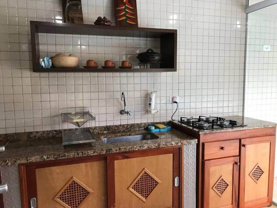 Cozinha da Casa dentro cond. Tenório.  Uma pia e um fogão estão lado a lado, abaixo deles um armário. Na parede acima da pia há uma prateleira com xícaras e outros objetos.