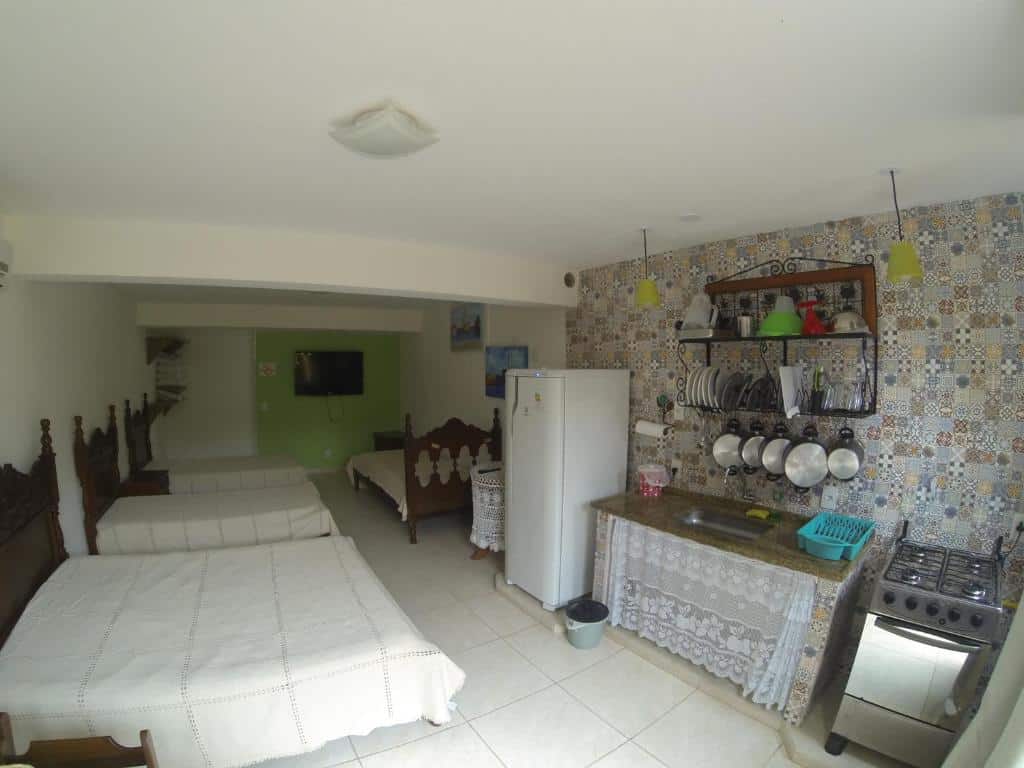Interior da Casa do Monge Apartments, um dos airbnb em Ilhabela. Na parede ao lado esquerdo há uma cama de casal e duas de solteiro, e elas encaram a cozinha compacta logo em frente. Há geladeira, pia com utensílios pendurados acima e fogão. Ao fundo do quarto há mais uma cama de casal e uma TV de tela plana.
