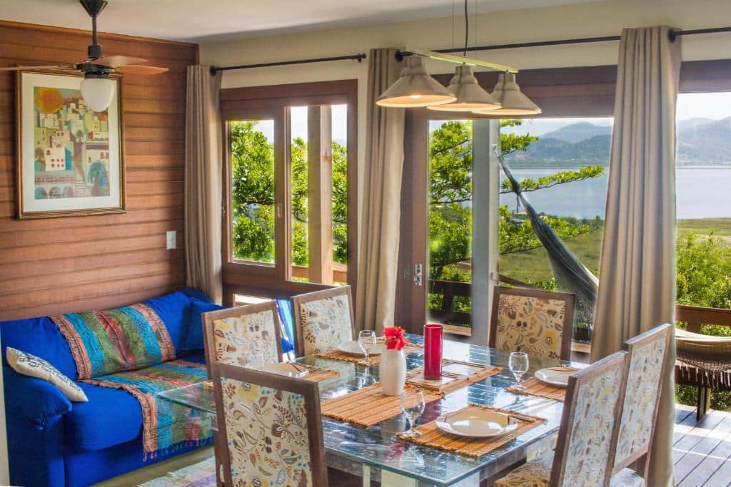 Sala da Casa em CondomÍnio Fechado com Vista para Lagoa. Uma mesa de jantar posta com seis cadeiras e utensílios está no centro do local. Na lateral esquerda está um sofá com almofadas e manta. Ao fundo há uma ampla porta de vidro que dá para a varanda com rede e vista do lago.