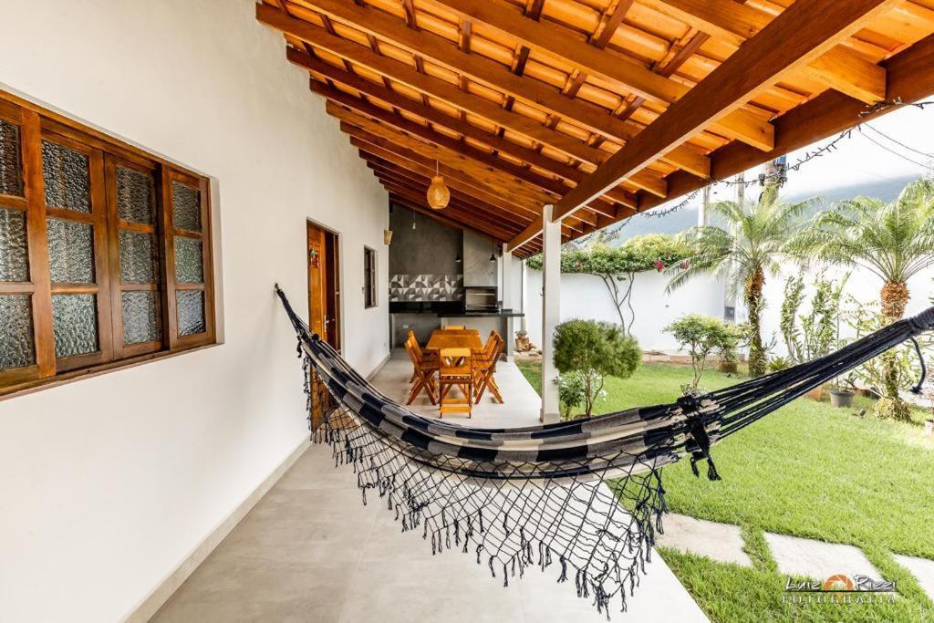 Área externa da Casa Flor. Do lado esquerdo estã a varanda, onde fica uma mesa de vários lugares e uma churrasqueira ao fundo. Mais a frente está uma rede. Do lado direito está o gramado.