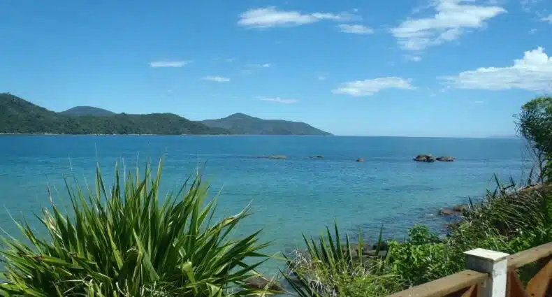 Vista da Casa frente ao mar c/ piscina para ilustrar post sobre airbnb na Praia da Enseada. Um apoio de madeira na frente com árvores e a vista do mar no fundo com montanhas.