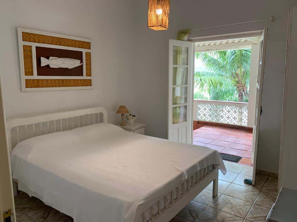 Quarto da Casa frente ao mar c/ piscina. Uma cama de casal do lado esquerdo e uma cômoda com abajur. No fundo uma porta aberta com acesso a varanda. Foto para ilustrar post sobre airbnb na Praia da Enseada.