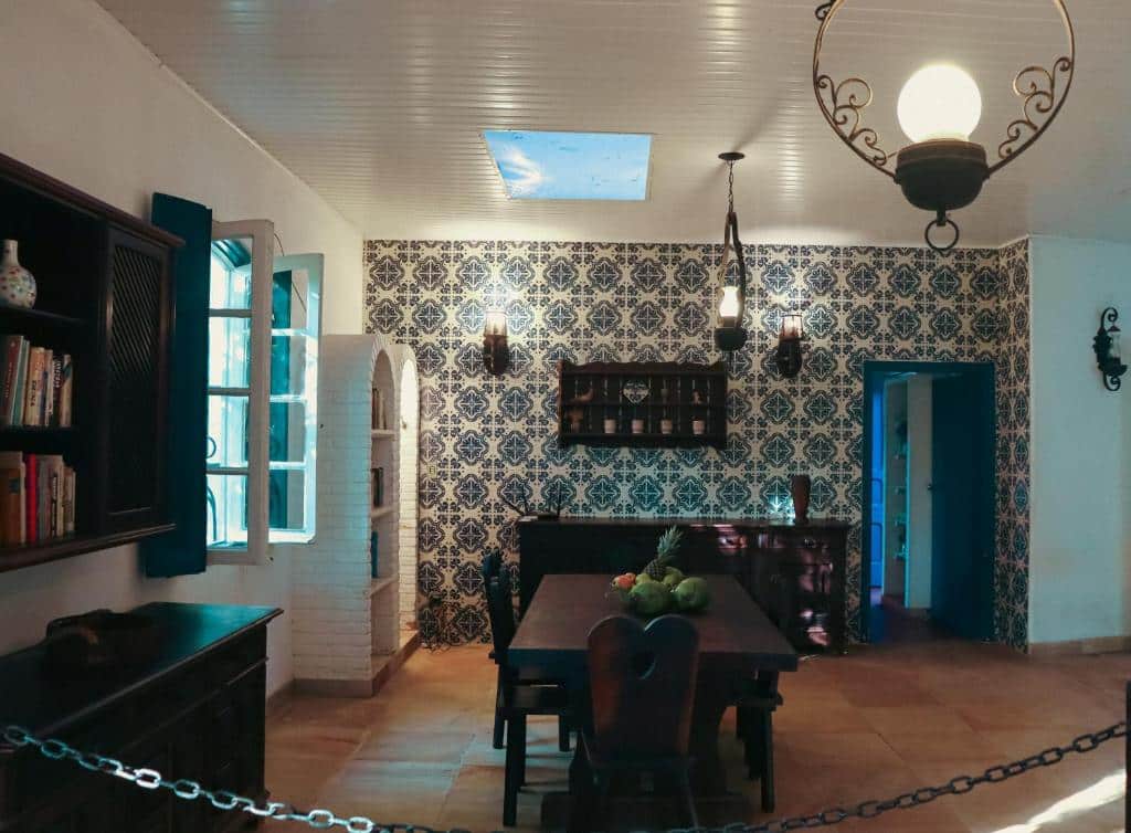 Sala de jantar da Casa na quadra da Praia do Tenório. Uma mesa está no meio do ambiente, de frente para um balcão encostado na parede do fundo. Ao seu lado esquerdo esá outro balcão, um armário encostado na parede e uma janela.