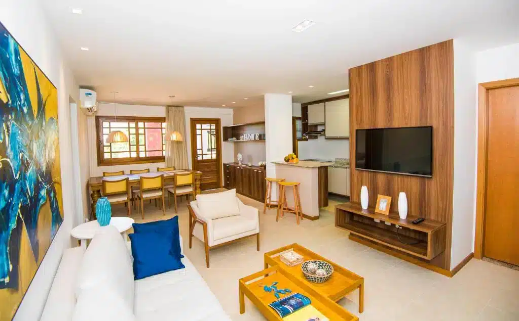 Sala e cozinha no Pé na areia - Furstberger. Em primeiro plano há uma sala espaçosa co m sofá à esquerda mesa de centro, poltrona e TV no centro em um painel de madeira. No fundo há uma cozinha completa em estilo americano e uma mesa de jantar com seis lugares.
