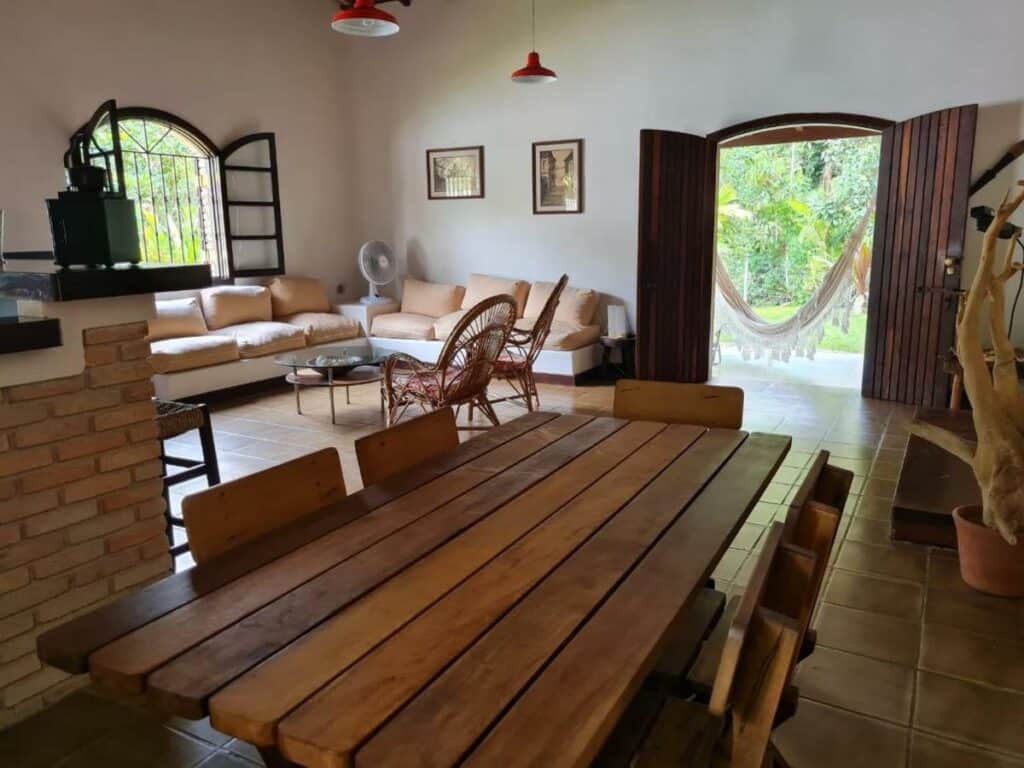 Sala de estar da Casa Praia Condomínio Prumirim Ubatuba. Em primeiro plano há uma grande mesa de madeira de vários lugares. Ao fundo está a porta e uma rede do lado de fora. Do outro lado estão os sofás, cadeiras e mesa de centro.