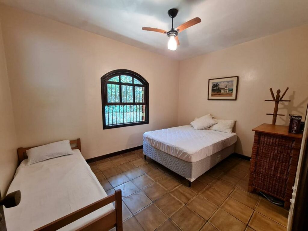 Quarto da Casa Praia Condomínio Prumirim Ubatuba. Uma cama de casal e uma cama de solteiro estão de frente uma para outra. Na parede há uma janela. Ao lado da cama de casal está um cabideiro e um móvel.