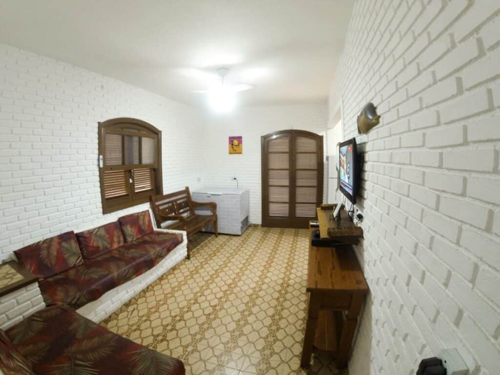 Sala da Casa Aconchegante a 200 mts da Praia da Maranduba. Do lado esquerdo estão dois sofás e um banco de madeira. De frente para eles, no lado esquerdo, está a tv. No fundo há um freezer encostado na parede e uma porta.