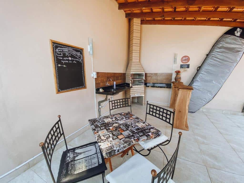 Área externa da Casa Ubatuba Vista para o Mar. Uma mesa com cadeiras de ferro, no fundo uma pia com bancada e uma churrasqueira. Do lado direito uma prancha em pé.