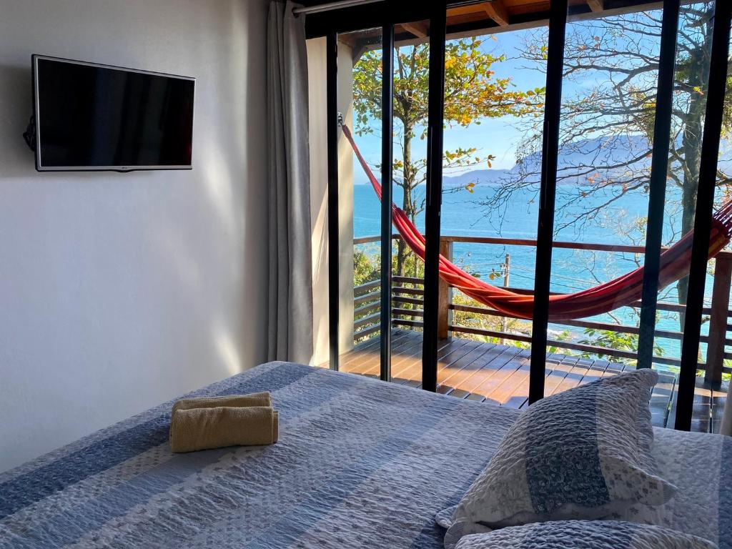 Quarto de uma das Casas Do Mar, um dos airbnb em Ilhabela. Uma cama de casal com duas toalhas dobradas aos pés está encostada na parede ao lado direito e encara uma TV na parede oposta. Ao lado direito da cama há uma extensa porta de vidro com cortinas que leva à varanda com rede e vista para o mar.