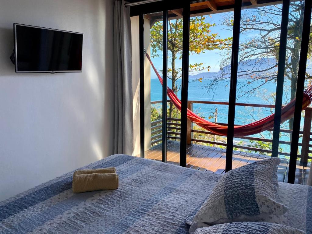 Quarto de uma das Casas Do Mar. Uma cama de casal com duas toalhas dobradas aos pés está encostada na parede ao lado direito e encara uma TV na parede oposta. Ao lado direito da cama há uma extensa porta de vidro com cortinas que leva à varanda com rede e vista para o mar.