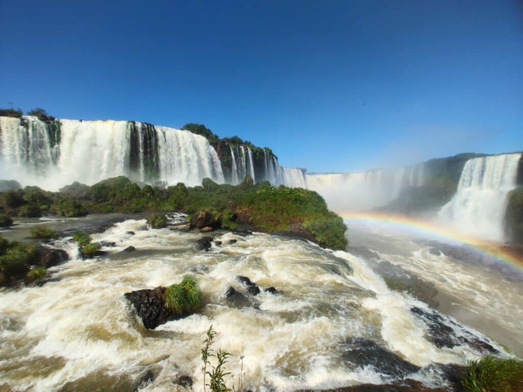 Imagem das Cataratas do Iguaçu com exuberantes quedas d´água, uma parte de plantas verdes e um lindo arco-íris entre as águas em movimento durante o céu azul.