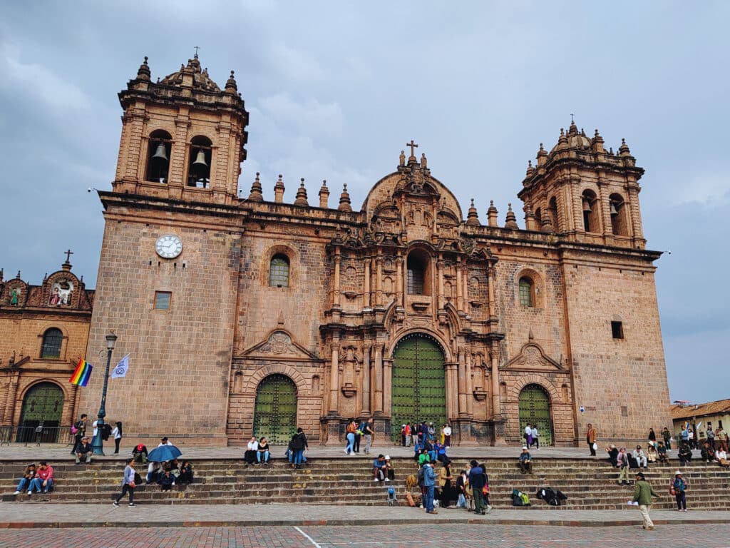 Catedral de Cusco vista de frente na plaza de armas. Uma construção antiga, com tijolos, duas torres de cada lado e três portas verdes na entrada, onde há uma grande escadaria